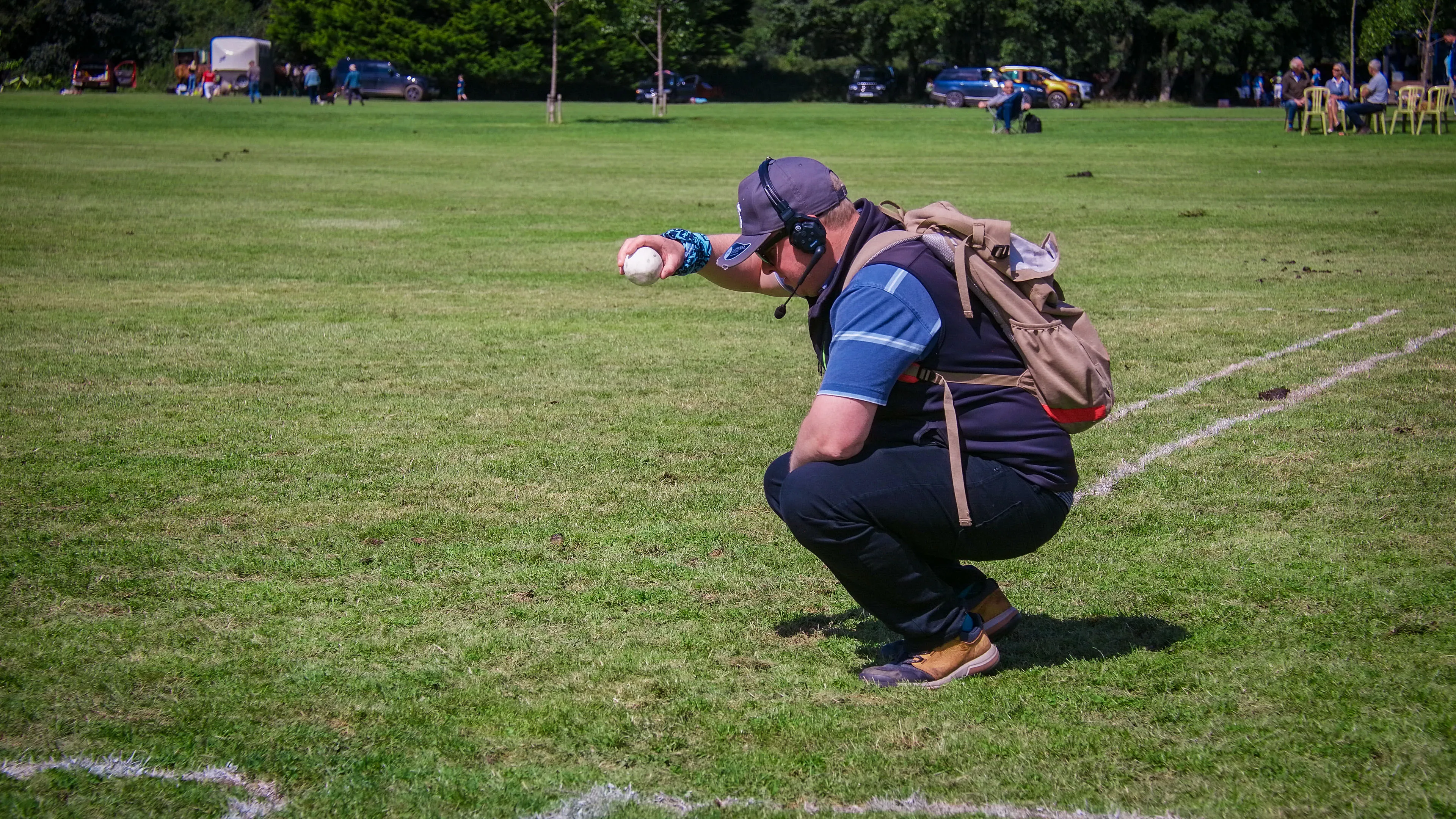A man squats on a grassy field, wearing a backpack and headset, ready to drop a ball. The scene conveys concentration.